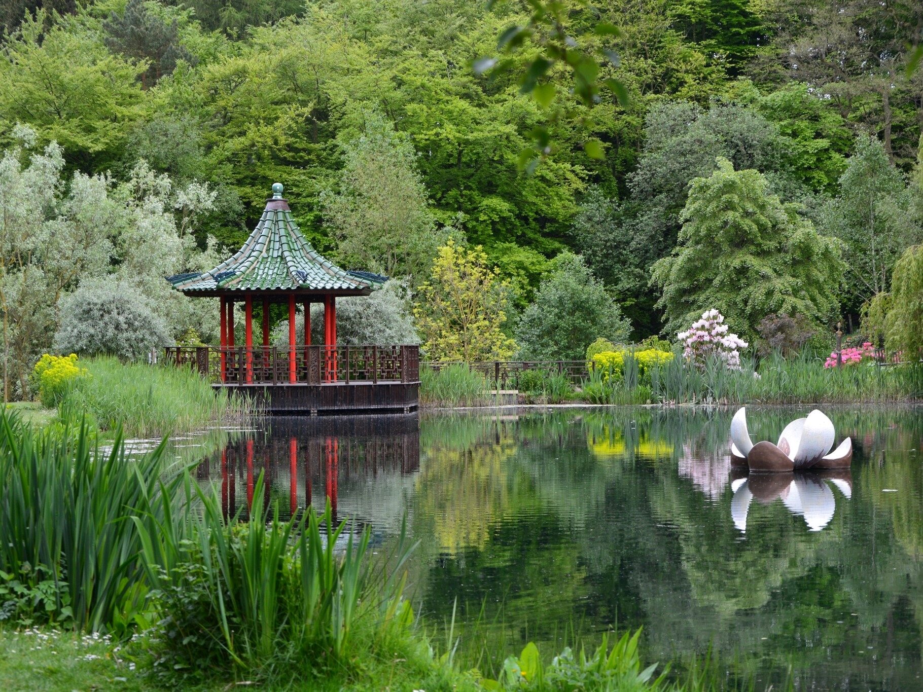 A lake with a large flower scuplture in it to the right. On the left is a red chinese style pagoda with a pointed roof. All around the edge of the lake and behind is a vast array of green trees and shrubs.