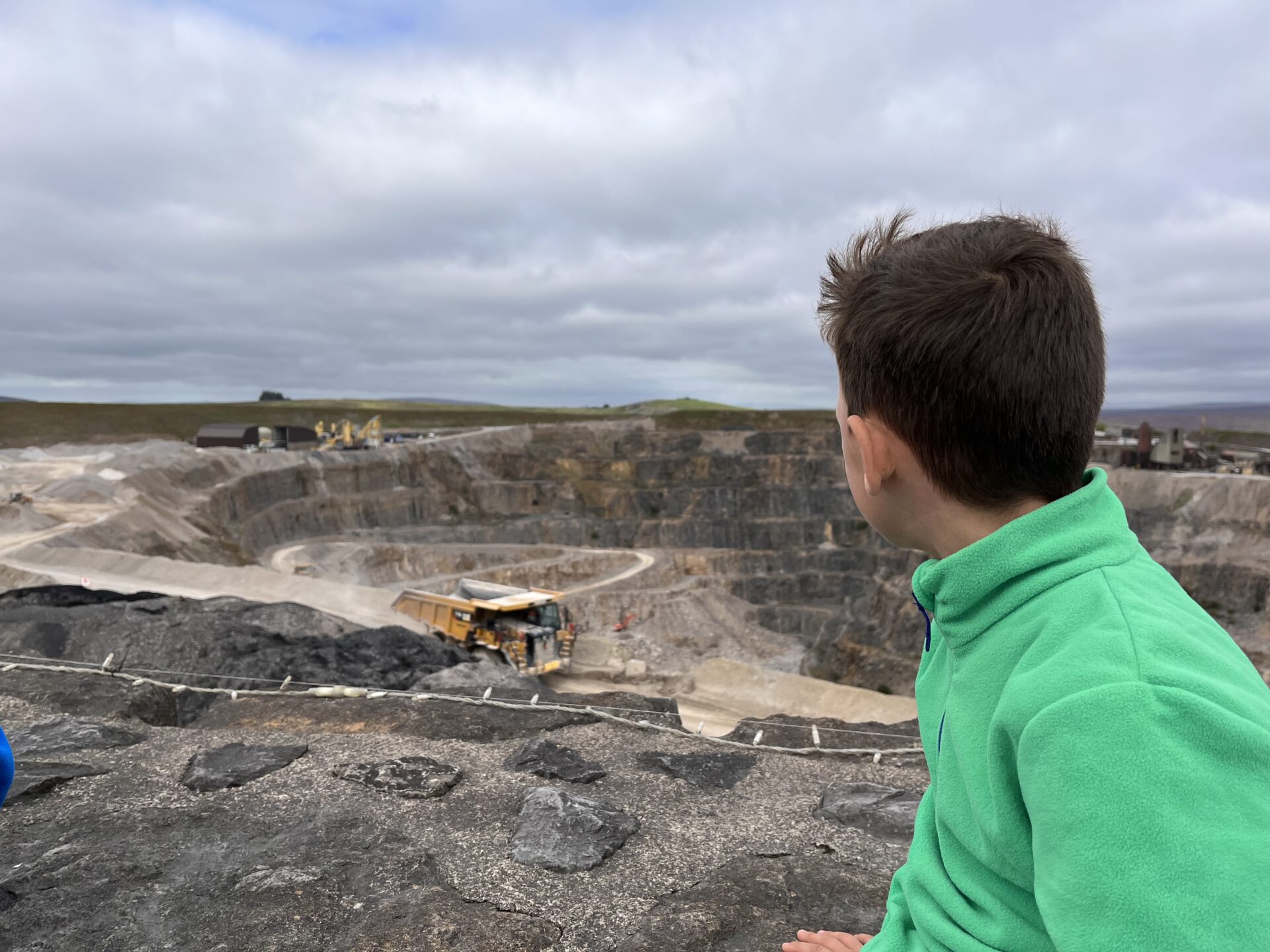 A view into Coldstones Quarry with a large yellow dumper truck driving past and a child wearing a green jumper looking into the quarry.