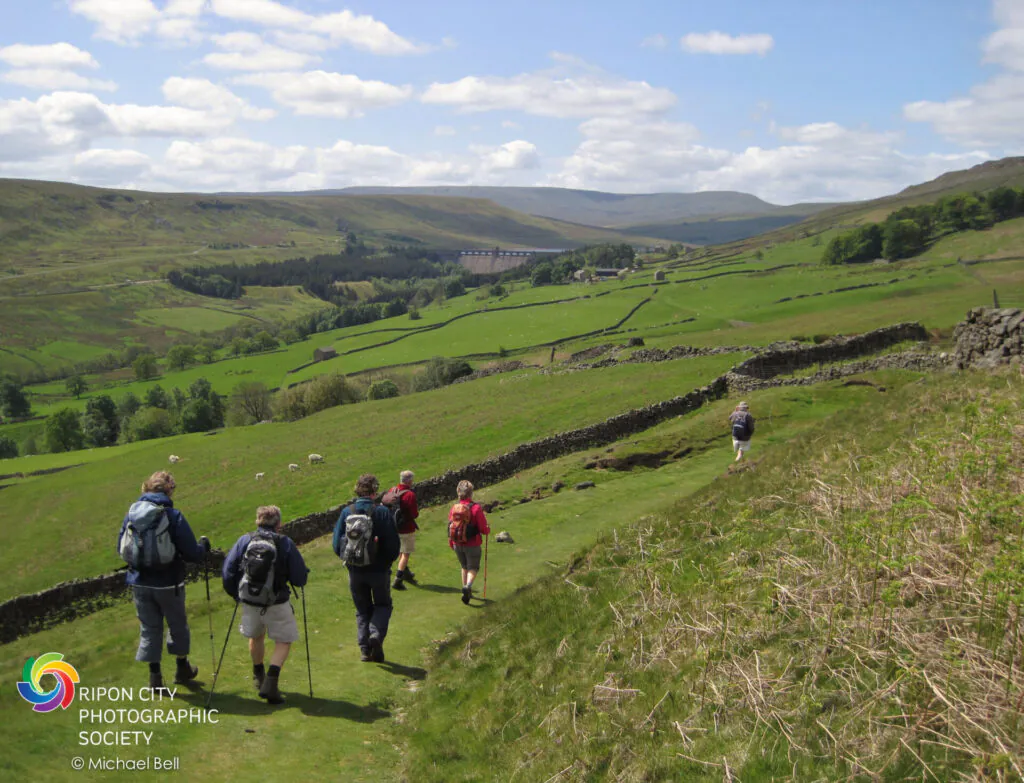 Blue sky and green fields with hills and Scar House Reservoir in the distance. A group of six walkers with backpacks and walking poles with their backs to the camera walking towards Scar House.